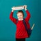 Cheerful thoughtful little school boy in school uniform with backpack and big pile of books standing against blue wall. School co