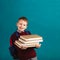 Cheerful thoughtful little school boy in school uniform with backpack and big pile of books standing against blue wall. School co