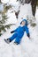 Cheerful toddler boy in blue overalls lies on snow