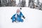 Cheerful toddler boy in blue overalls sits on snow