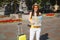 Cheerful traveler tourist woman in hat, yellow clothes, orange heart glasses with suitcase hold city map in city outdoor