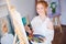 Cheerful woman artist standing and painting picture in workshop