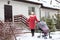 Cheerful woman in red warm long jacket standing next to townhouse porch and entrance at winter season, walking with baby buggy