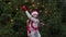 Cheerful woman in Santa hat with red scarf and mittens is happy and dancing on a background of Christmas tree in