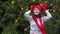 Cheerful woman in santa hat with red scarf and mittens is happy and jumping against the background of Christmas tree in