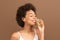 Cheerful young african american curly woman with perfect skin in white top sniffing bottle of scent