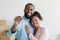 Cheerful young african american husband hugs wife and shows keys in room with cardboard boxes