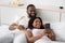 Cheerful young african american husband hugs wife with smartphone on white bed in bedroom interior