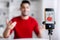 Cheerful young arab man with beard gesticulates and shoots video for blog at phone in home interior, blurred