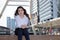 Cheerful young Asian businesswoman sitting on staircase after work and positive look at urban building background.