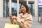 Cheerful young Asian woman in winter outfit holding gift bags, using cellphone, shopping online via mobile app outdoors