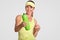 Cheerful young athletic woman with charming smile, suggests you to drink, holds green bottle with water, dressed in sportswear, is