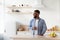 Cheerful young bearded black guy with glass of clean water in hand in kitchen interior looks at window