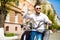 Cheerful young bearded man in helmet is sitting on scooter and say hello on city street