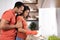 Cheerful young black male hugs wife in rubber gloves, lady washes dishes in kitchen interior, profile