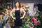 Cheerful young florist stand inside at black table. She hold hands on hips and smile on camera. Young woman stand inside