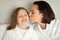 Cheerful young lady kissing on cheek little caucasian girl in bathrobe, relaxing on white bed, enjoy spare time