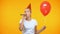 Cheerful young lady in party hat blowing horn, holding red balloon, birthday