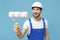 Cheerful young man in coveralls protective helmet hardhat hold paint roller isolated on pastel blue background in studio