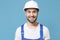 Cheerful young man in coveralls protective helmet hardhat isolated on blue wall background studio portrait. Instruments