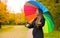 Cheerful young woman with colorful umbrella checking for rain in city park