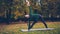 Cheerful young woman is doing yoga balancing exercises during individual practice in city park standing on mat and