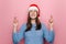 Cheerful young woman stands with fingers crossed, awaits miracle happen, wears Christmas hat and cozy sweater