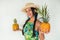A cheerful young woman in a straw hat holding whole pineapple fruits, looking at the camera, smiling