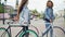 Cheerful young women are talking while walking with bikes along beautiful street with fountain and beautiful buildings