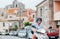 Cheerfully smiling man in helmet and sunglasses fast riding the moto scooter on the Sicilian old town streets with parked cars in