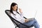 Cheery woman in headphones having online video call on laptop, sitting on chair against white wall, full length