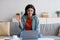 Cheery young Arab female wearing headphones, using laptop pc, working or studying online, waving at webcam indoors