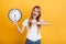 Cheery young blonde woman posing isolated over yellow wall background dressed in white casual t-shirt holding clock pointing