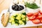 Cheese in brown saucer, bowls with dill, black olives, pieces of cucumbers, tomato, knife on cutting board on table