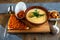 Cheese soup mashed in a clay plate is on a tray. Next to him is a piece of bread, a spoon and toast in a plate