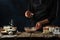 The chef in black apron pours cinnamon into glass bowl with dough for cooking waffle on rustic wooden table with ingredients on