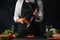 The chef in black apron shredding carrots with grater on black chopped board at professional restaurant kitchen on dark blue