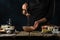 The chef in black apron testing by ladle dough for cooking waffle on rustic wooden table with ingredients on dark blue background