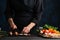 The chef in black uniform cuts with knife mushrooms on the black chopping board at the professional kitchen on dark blue