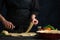 The chef in black uniform cuts with round knife dough for cooking lasagna. Traditional recipe. Italian cuisine. Dark background.
