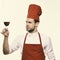 Chef in burgundy uniform with glass of red wine. Cook with satisfied face looks at wineglass. Italian drink concept.