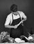 Chef with busy face holds blue and metal knives on dark green background. Man in cook hat and apron sharpens blades.