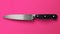 A chef\\\'s knife with a sleek blade and black handle lies against a striking pink background