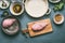 Chicken breast on cutting board with cooking ingredients bowls on gray background, top view