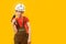 Child as builder. Boy in white helmet, protective glasses wears work jumpsuit and holds drill. Isolated on yellow background