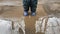 A child in blue countries and rubber boots stands in a muddy puddle, then squats and jumps, splashes of water flying in