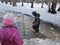 A child in blue waterproof pants, a jacket and a hat stands in a deep puddle. A girl in a pink jacket is looking