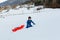 child in blue winter clothes climbing up the snowy hill pulling the red sled