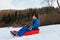 Child in blue winter clothes sliding down the snowy hill in red sled