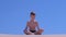 Child boy is meditating in lotus pose on sandy beach on blue sky background.